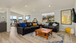 A spacious living room with a black leather sofa, a wooden coffee table, and a large rug. There are two windows with views of the outdoors. - Hood Canal Beach house, Low bank waterfront,  enjoy nature from your deck (Belfair)