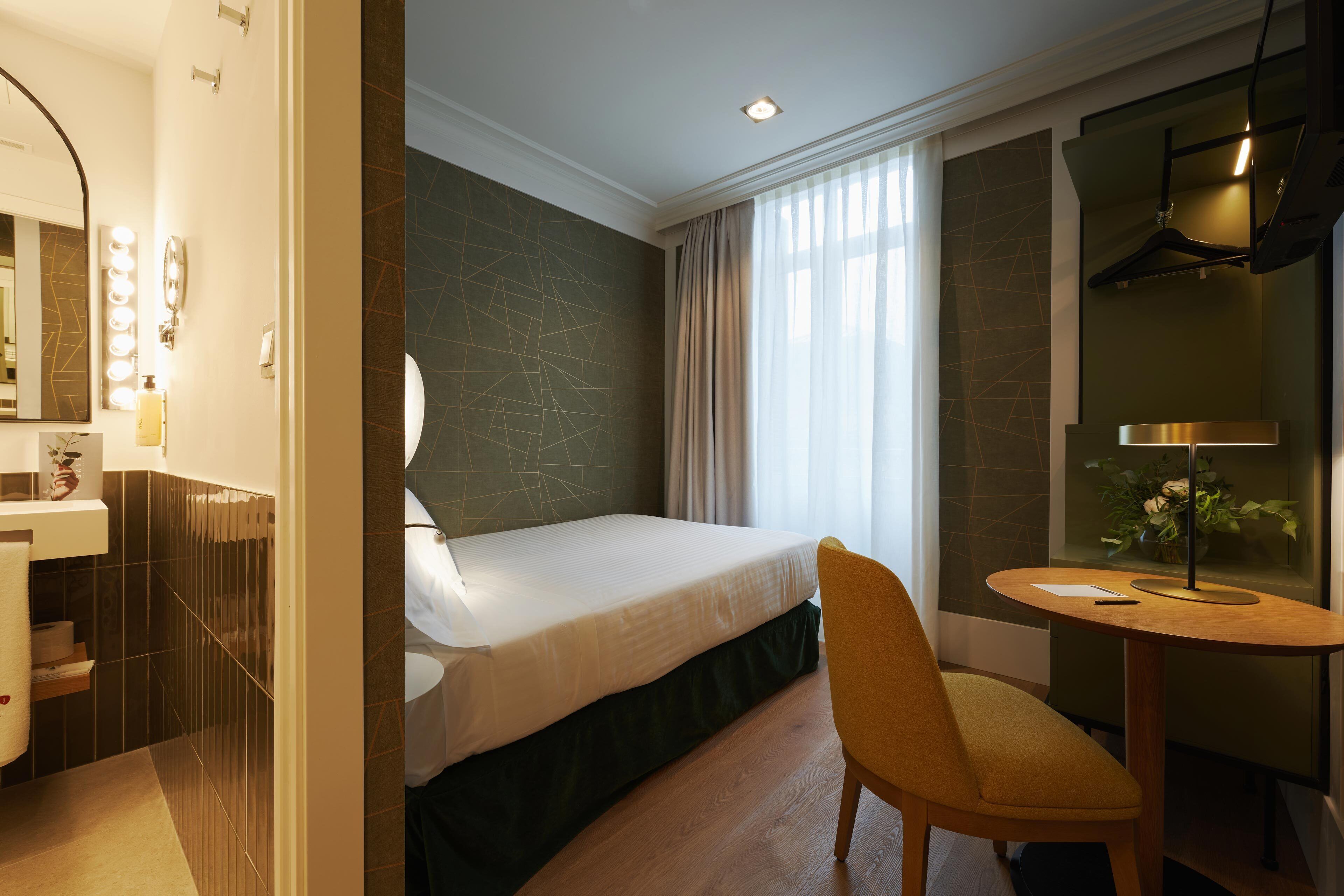 A hotel room with a bed, a chair, a desk, a mirror, and a bathroom with a sink and mirror.
