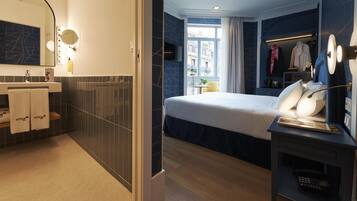 Una habitación de hotel moderna con una cama grande, una mesita de noche con lámpara, un tocador con espejo e iluminación, y vistas a la ciudad a través de la ventana.