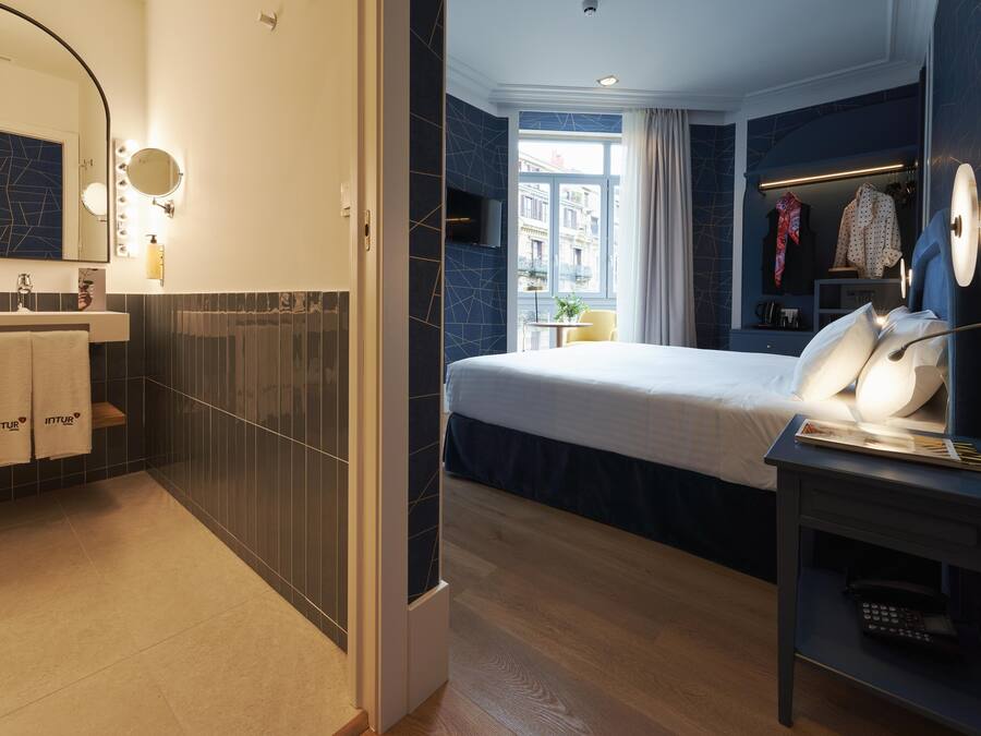 Una habitación de hotel moderna con una cama grande, una mesita de noche con lámpara, un tocador con espejo e iluminación, y vistas a la ciudad a través de la ventana.