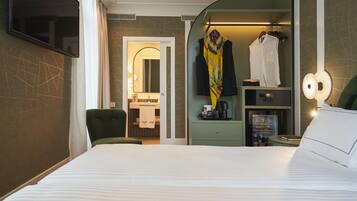 Una habitación de hotel con una cama, un sillón verde, una tocador con espejo y un perchero con ropa colgada.
