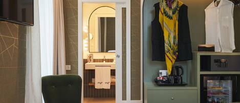 Habitación de hotel con un sillón verde, una cama, un minibar y un perchero con una prenda amarilla.