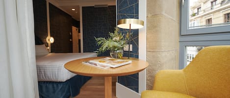 Habitación de hotel con cama, una mesa redonda de madera con una revista, un sillón amarillo, una lámpara de pie y vistas a edificios a través de una ventana.