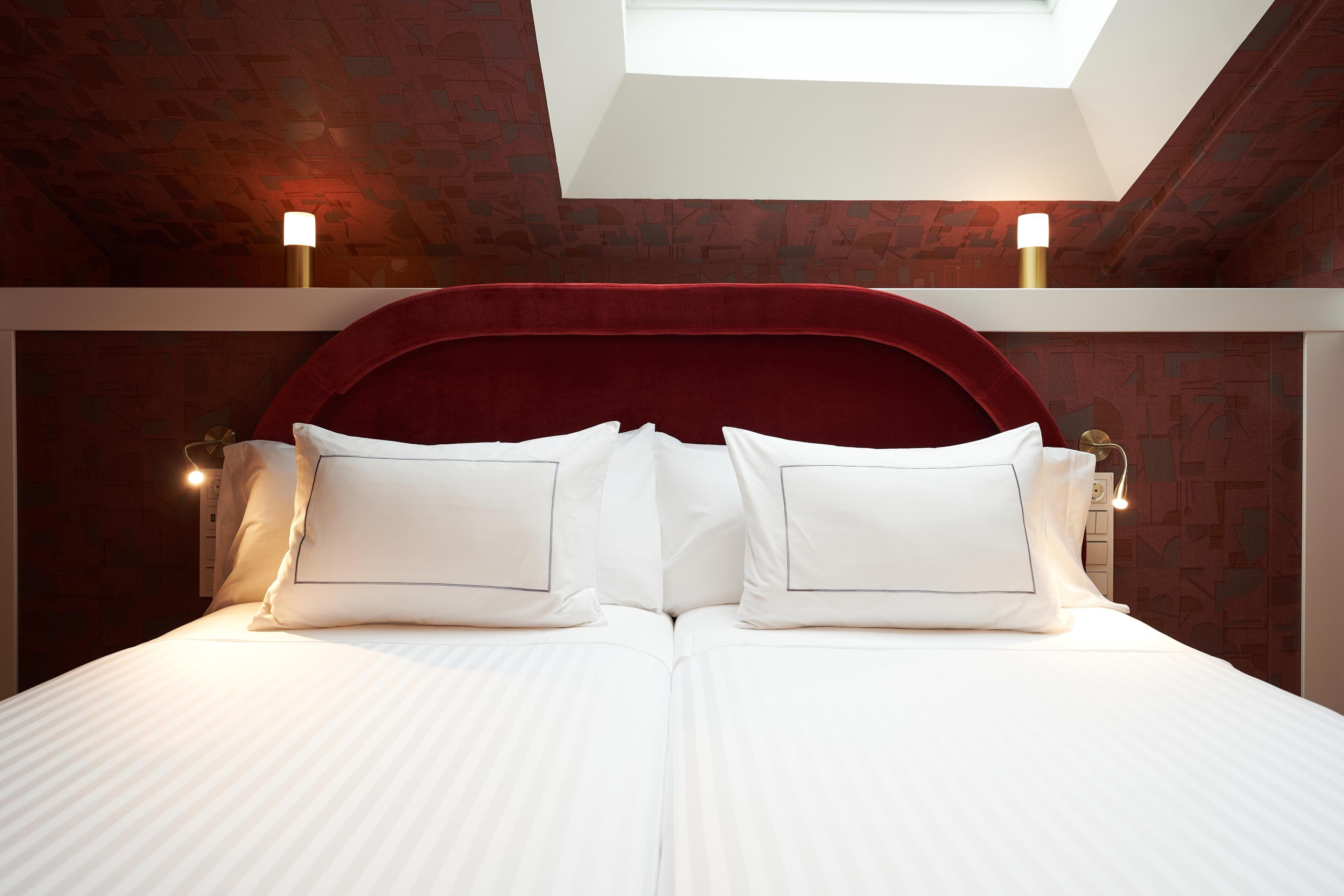 Un lit avec du linge de lit blanc et une tête de lit rouge, deux lampes de chevet et une lucarne au-dessus.