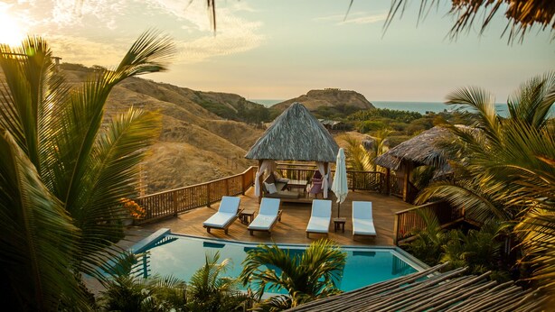 A poolside area with lounge chairs, a thatched-roof hut, and a view of the ocean and hills. - Villa del Sol Mancora (Máncora)