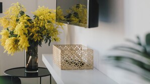 A modern living room with a vase of yellow flowers, a flat-screen TV, a woven basket, and a decorative box on a white shelf.