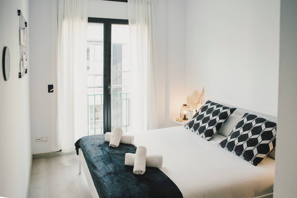 Habitación de hotel con cama, ventana con cortinas, mesita de noche con lámpara y vista al exterior.