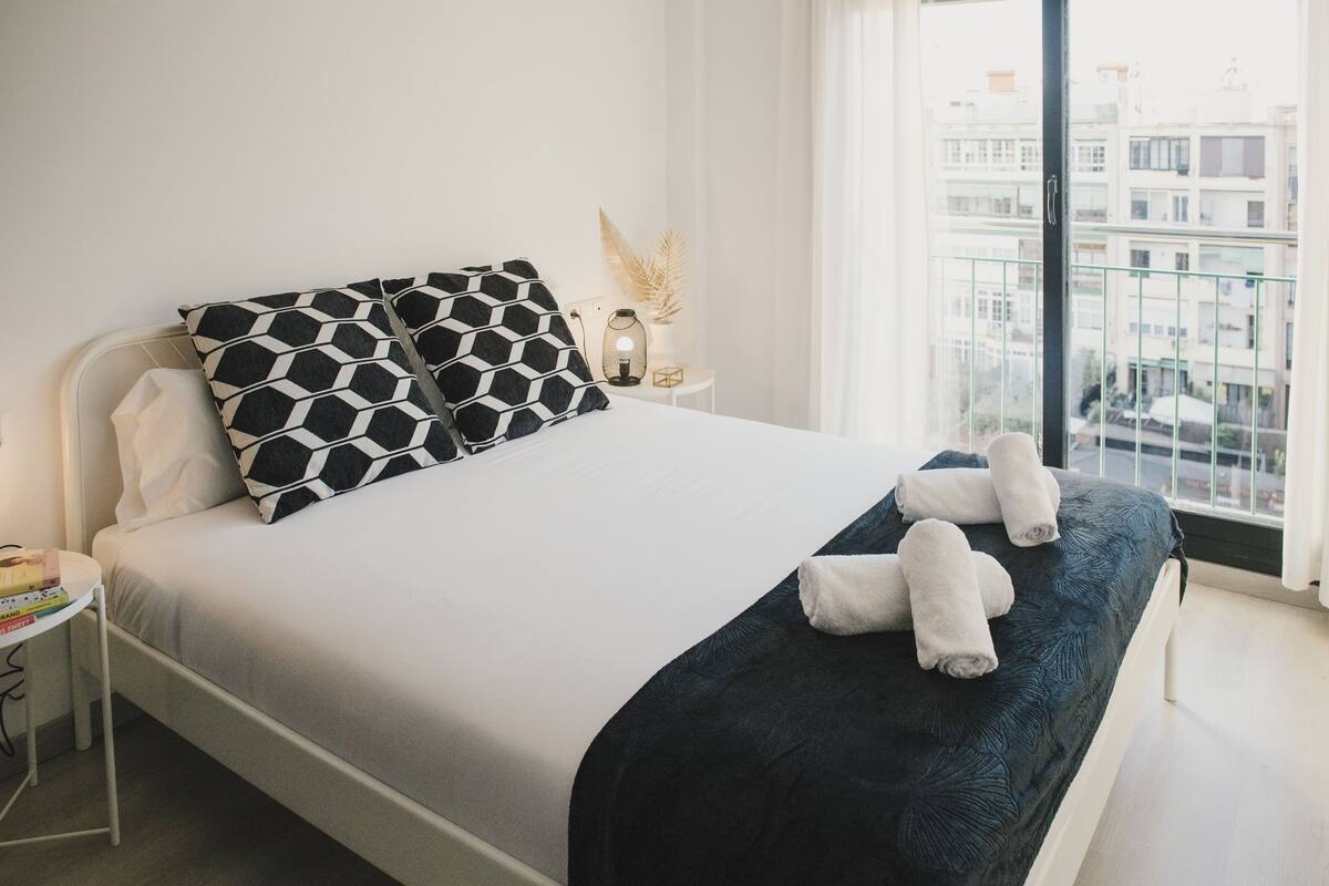 Una cama bien hecha con una almohada de diseño blanco y negro, una manta azul oscuro y dos toallas sobre la cama. Hay una mesita de noche con una lámpara y una planta decorativa. La habitación tiene un ventanal con vistas a edificios.