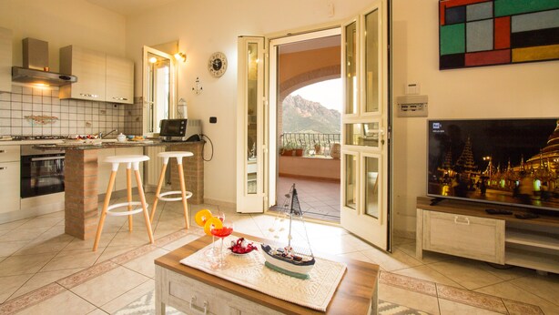 A modern kitchen with a dining area, a TV, and a view of mountains through an open door. - Villa Calu' - Wonderful villa in Sardinia with swimming pool and sea view (Tertenia)