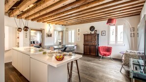 A spacious kitchen with a large island, wooden beams on the ceiling, and a red chair in the living area. - Exclusive apartment in the former Ospizio delle Donzelle, near Biennale Gardens (Venezia)