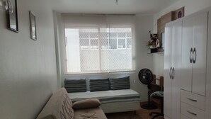 Een kleine, goed verlichte kamer met een bank, een zitje, een ventilator en een garderobe.