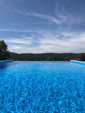 Une piscine d’un bleu éclatant, avec vue sur une colline boisée et quelques bâtiments visibles au loin.