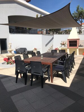 Une terrasse extérieure avec une longue table en bois, des chaises noires, une fontaine en pierre et un four en briques.