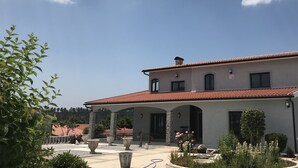 Un bâtiment à deux étages, doté d’un toit recouvert de tuiles rouges, d’un patio abrité et d’un jardin aménagé avec diverses plantes et une fontaine.