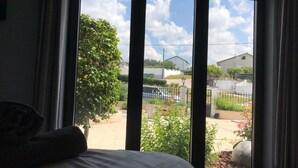 Une chambre avec une grande fenêtre, un lit et une vue sur un jardin et des bâtiments à l’extérieur.