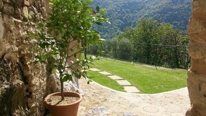 Un muro de piedra con un árbol en maceta, un patio empedrado y vistas a un área verde con una valla y montañas al fondo.