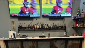 Un espace bar équipé de deux téléviseurs diffusant des matchs de sport, d’un comptoir en bois et d’un tuyau vert posé sur le sol.