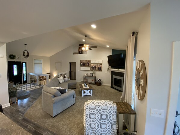 A spacious living room with a grey sofa, a fireplace, a television, and a patterned ottoman. - EAA 4 Bedroom House, Golf Simulator and Large Outdoor Patio. (Winneconne)