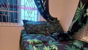 A bedroom with a bed covered in a green leaf-patterned bedspread, a window with metal bars, and a curtain with a leaf design. -  holiday stay, 2 bed 1 bathroom  (Port Morant)