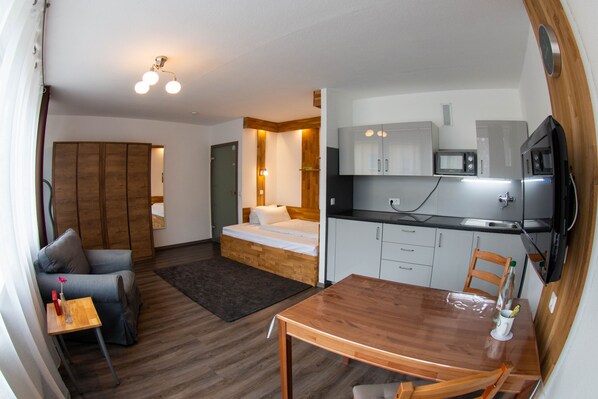 A compact room with a bed, a small sofa, a wooden dining table, and a kitchenette.