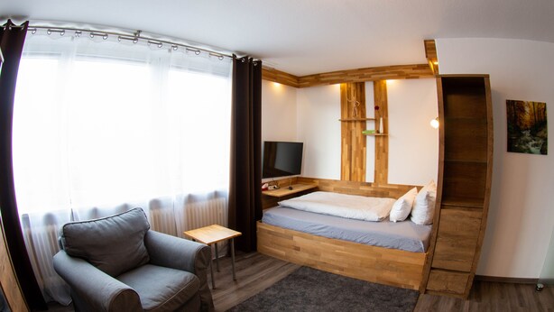 A bedroom with a bed, a TV, a small table, a chair, and a window with curtains. - LuPartment-Studio-Apartment-Stadtmitte (Ludwigshafen)