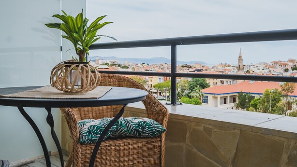 Een balkon met een rieten stoel, een klein tafeltje en een bloempot. Het balkon heeft uitzicht op een stadsgezicht met gebouwen en een berg op de achtergrond.