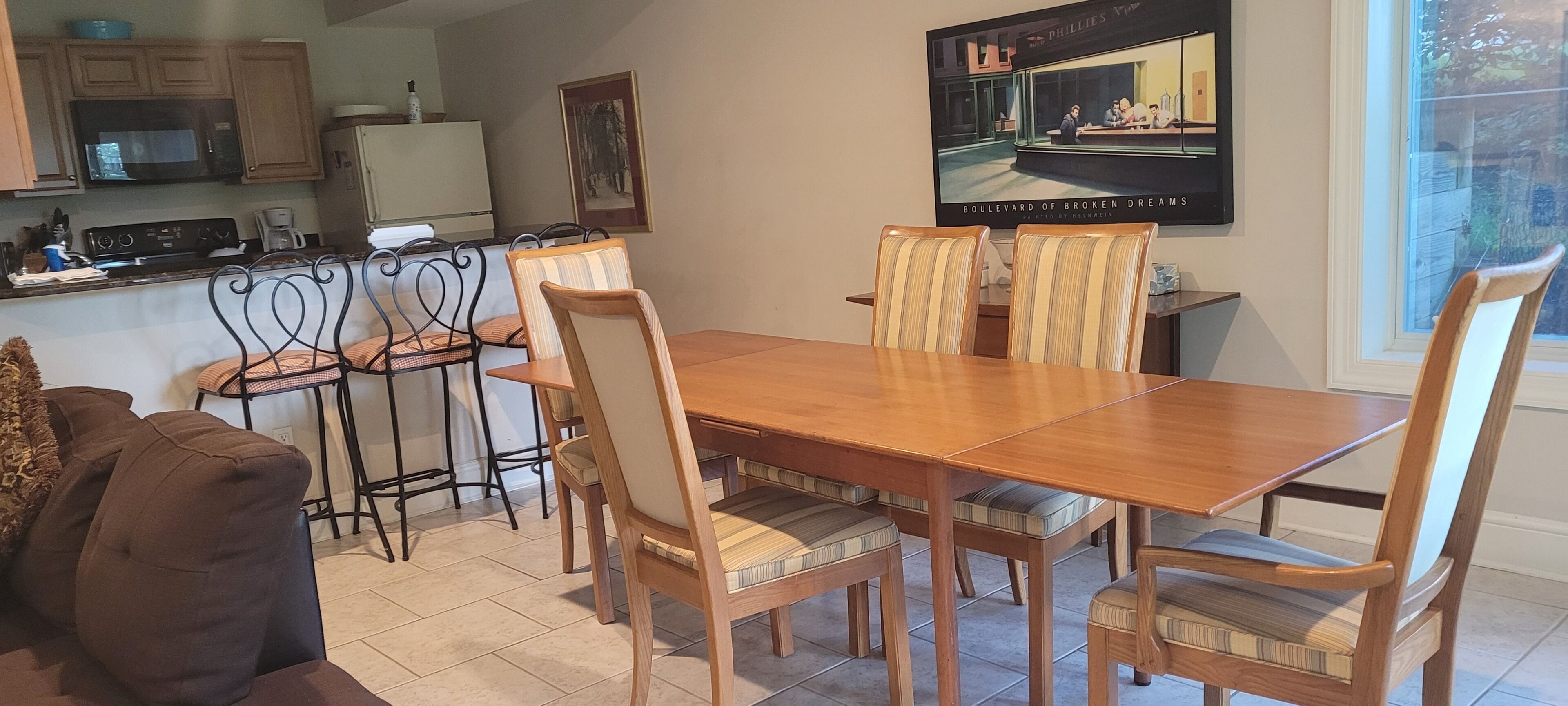 Een eethoek met een houten tafel en stoelen, een keuken op de achtergrond en een ingelijst schilderij aan de muur.