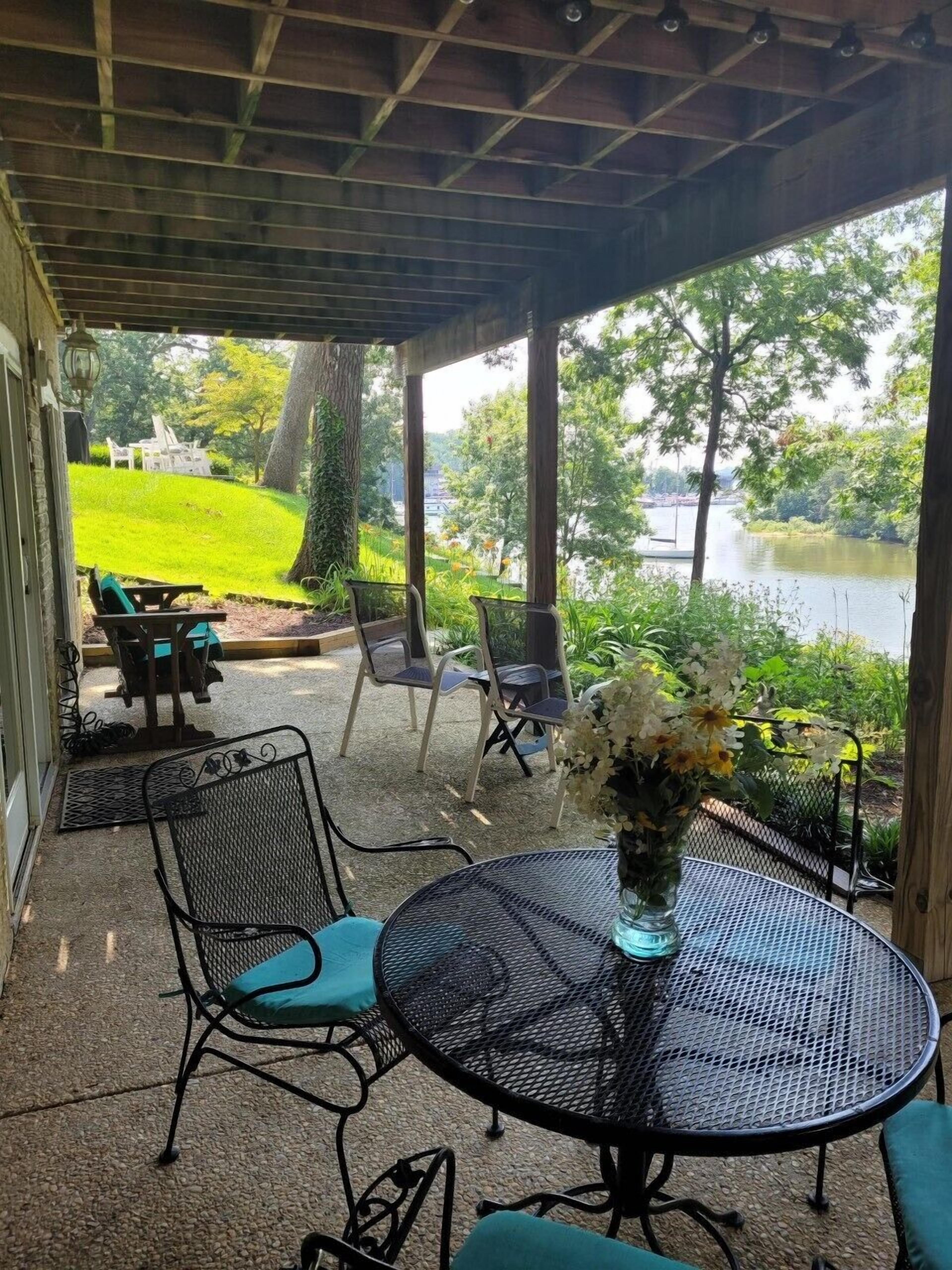 Une terrasse couverte avec une table ronde et des chaises, un vase de fleurs et une vue sur un lac et la verdure environnante.