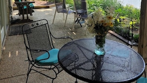En overdækket terrasse med et rundt bord og stole, en blomstervase og udsigt til et søområde og grønt.