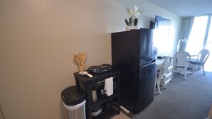 A compact kitchen with a black refrigerator, a small countertop with a stove, a trash bin, and a white chair in the dining area. - Island Colony 715 Charming Studio with Pool View (Honolulu)