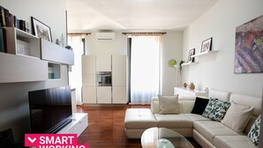 Un salon moderne avec un canapé, une table basse et un téléviseur. Il y a un coin cuisine avec des placards et un four. La pièce est dotée d’une grande fenêtre avec des rideaux.