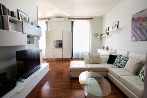 A modern living room with a flat-screen TV, a glass coffee table, a beige sofa, and a kitchen area with cabinets and an oven.