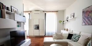 Ein modernes Wohnzimmer mit Sofa, Couchtisch und Fernseher. Es gibt eine Küchenzeile mit Schränken und einem Backofen. Das Zimmer hat ein großes Fenster mit Vorhängen.