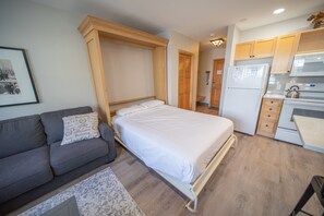 Una habitación pequeña con cama, sofá, kitchenette y una puerta que da a otra estancia.