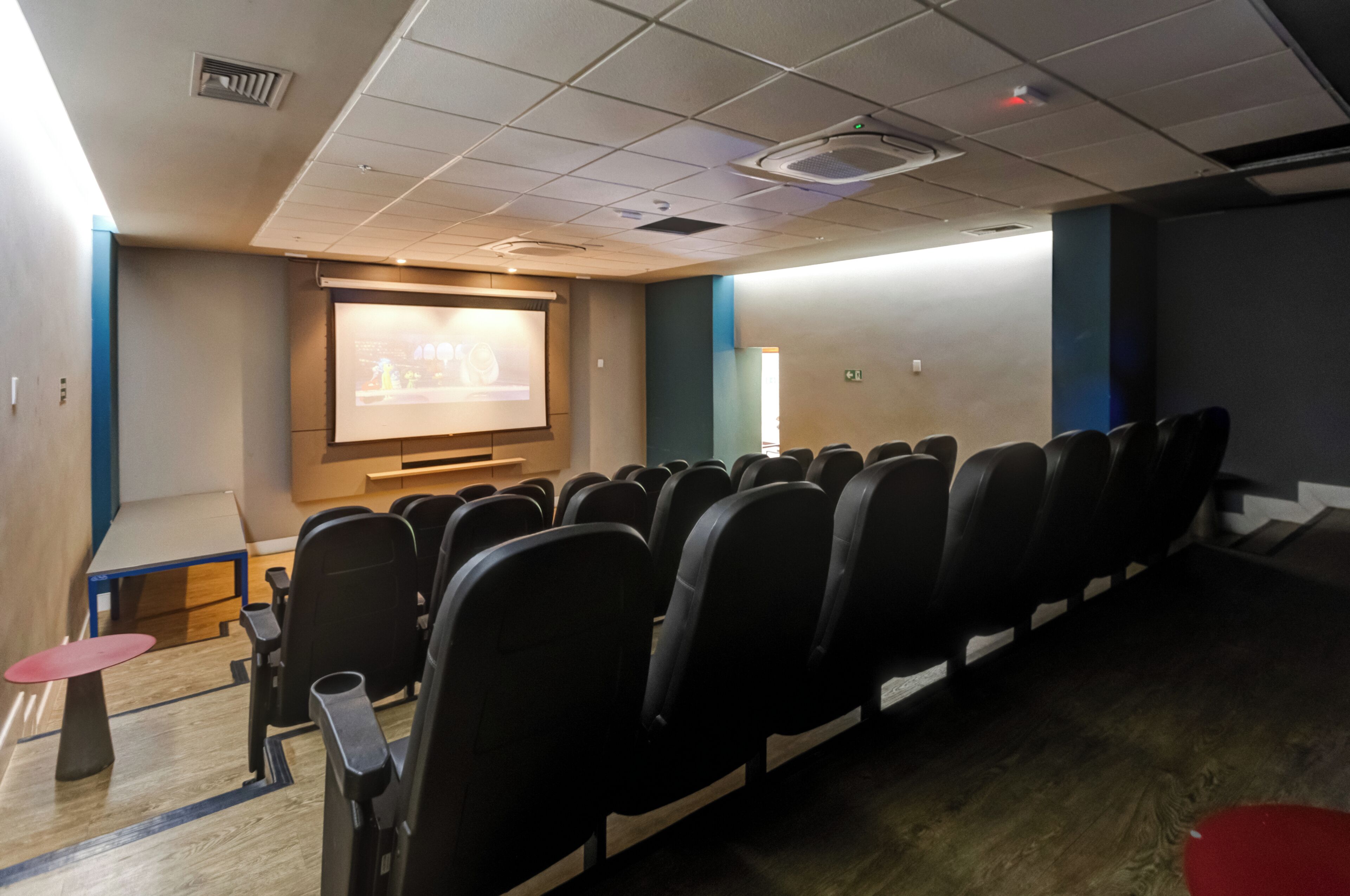 Une salle de conférence avec des rangées de chaises, un écran de projection et une petite table surmontée d’un tabouret rouge.
