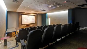 Una sala conferenze con file di sedie, uno schermo per proiettore e un piccolo tavolo con uno sgabello rosso.