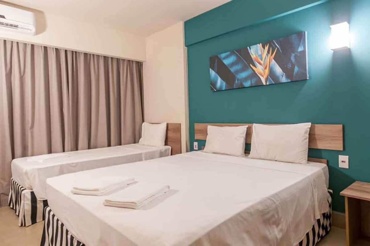 Een hotelkamer met twee bedden, een groot raam met gordijnen, een wandairconditioner en een schilderij boven de bedden.