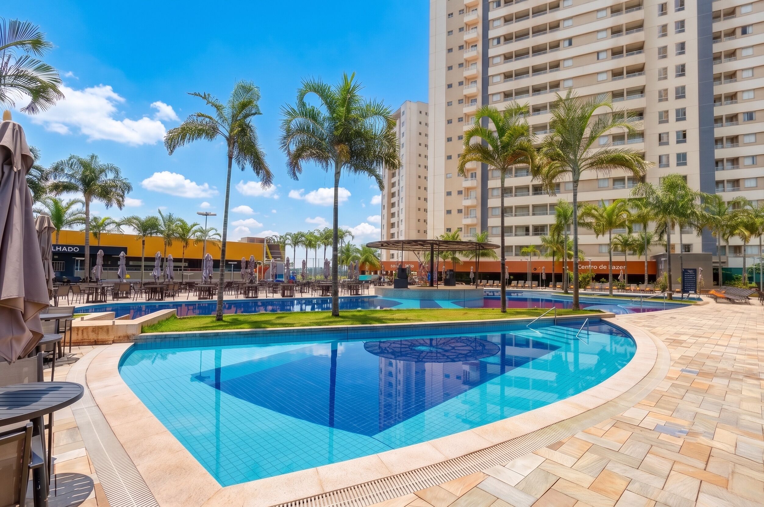 A swimming pool with a tiled deck, surrounded by palm trees and a multi-story building in the background.