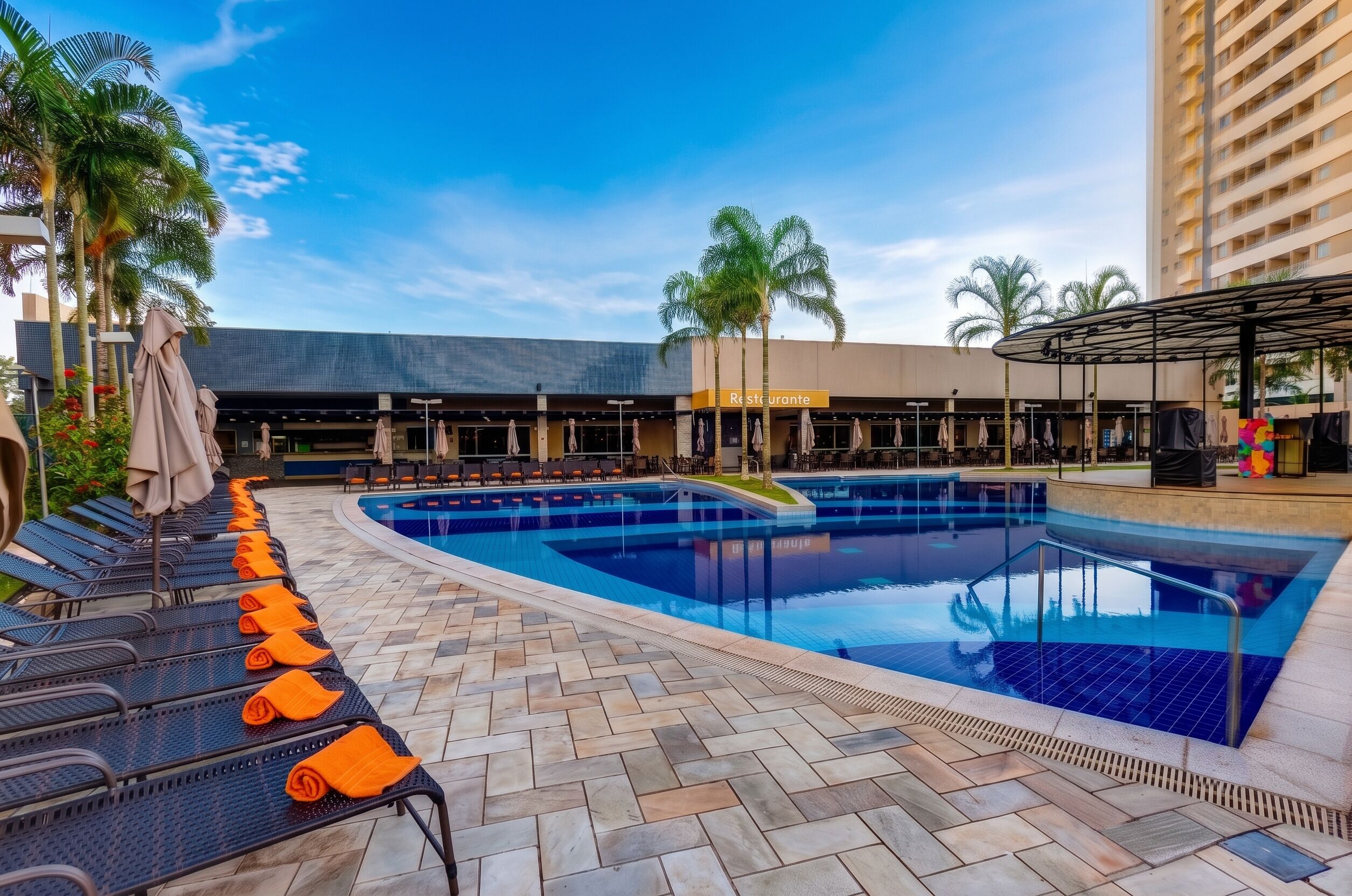 A pool area with lounge chairs and umbrellas, a building labeled 'Restaurante' in the background, and palm trees.