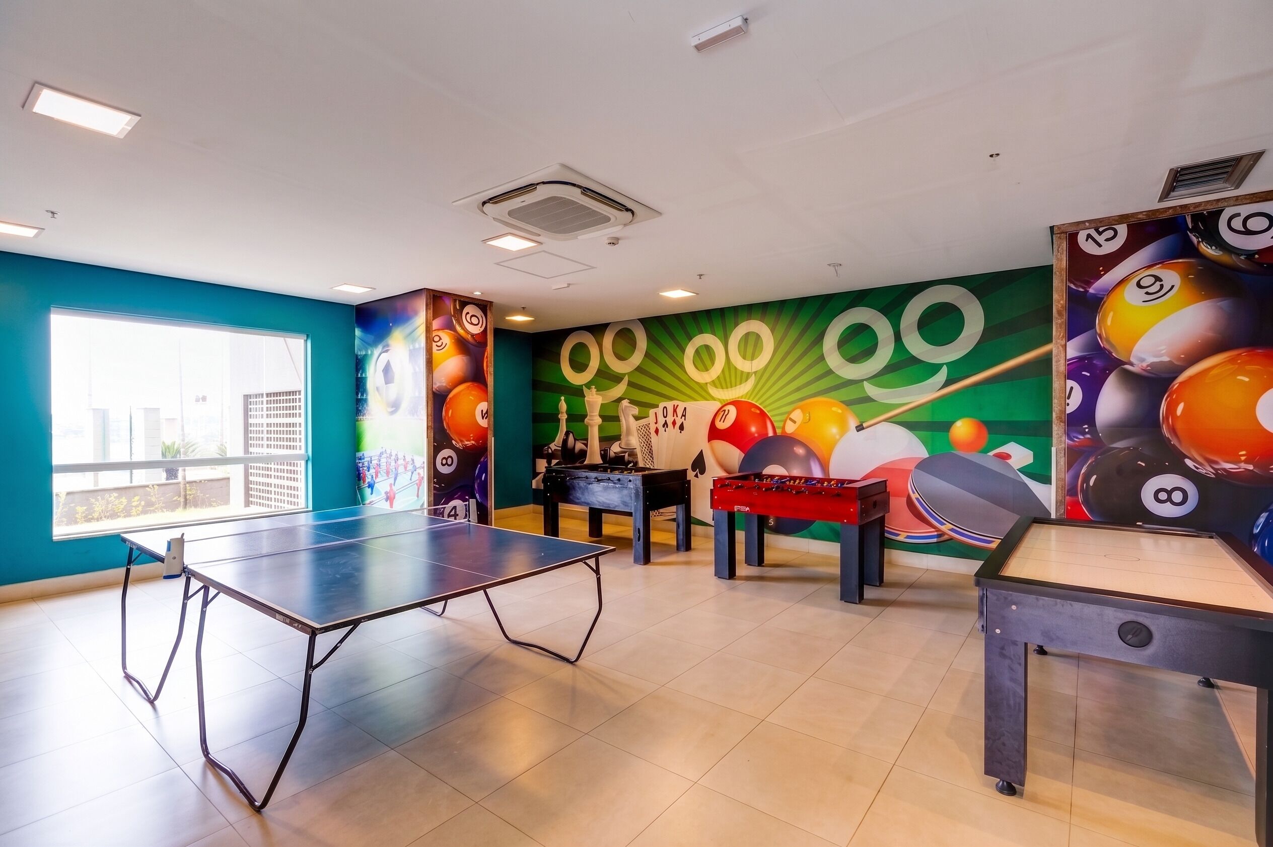 A room with a ping pong table, foosball table, and a billiards table. There are large windows on one side.