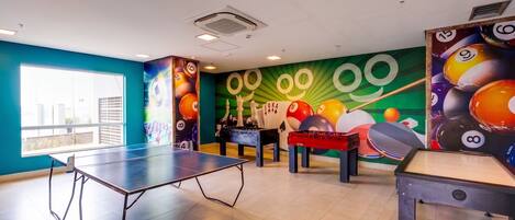 A room with a ping pong table, foosball table, and a billiards table. There are large windows on one side.
