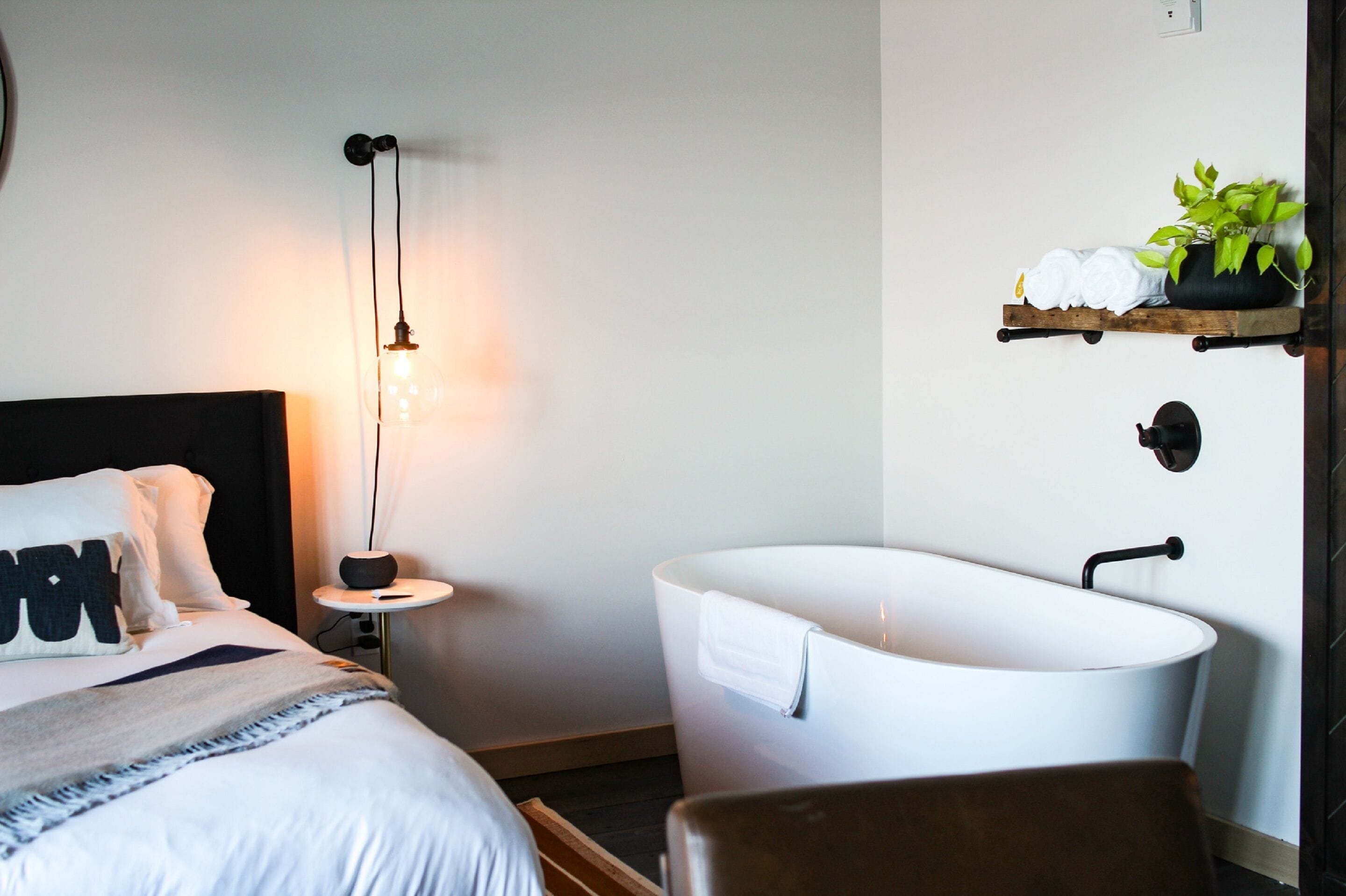 Un dormitorio moderno con una cama grande, una mesita de noche con lámpara, una bañera blanca y un estante con plantas.