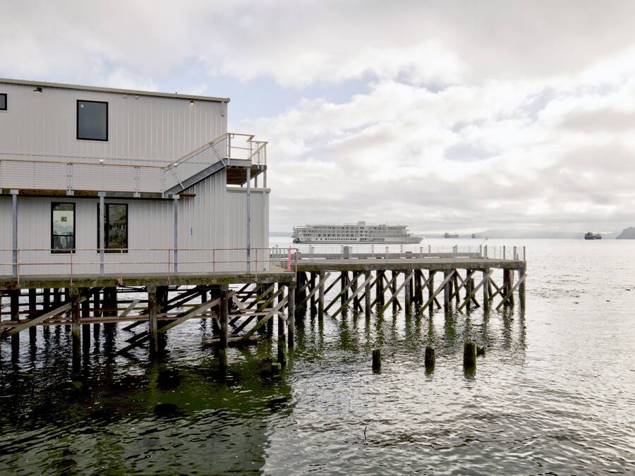 Een gebouw aan het water met een steiger die in het water uitloopt, een groot schip op de achtergrond en een bewolkte lucht.
