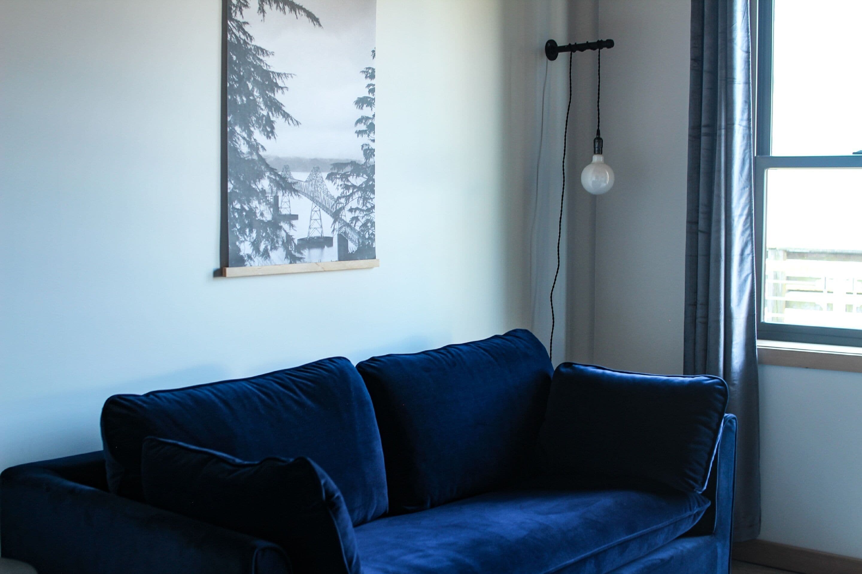 A room with a blue sofa, a framed picture on the wall, a hanging light bulb, and a window with curtains.