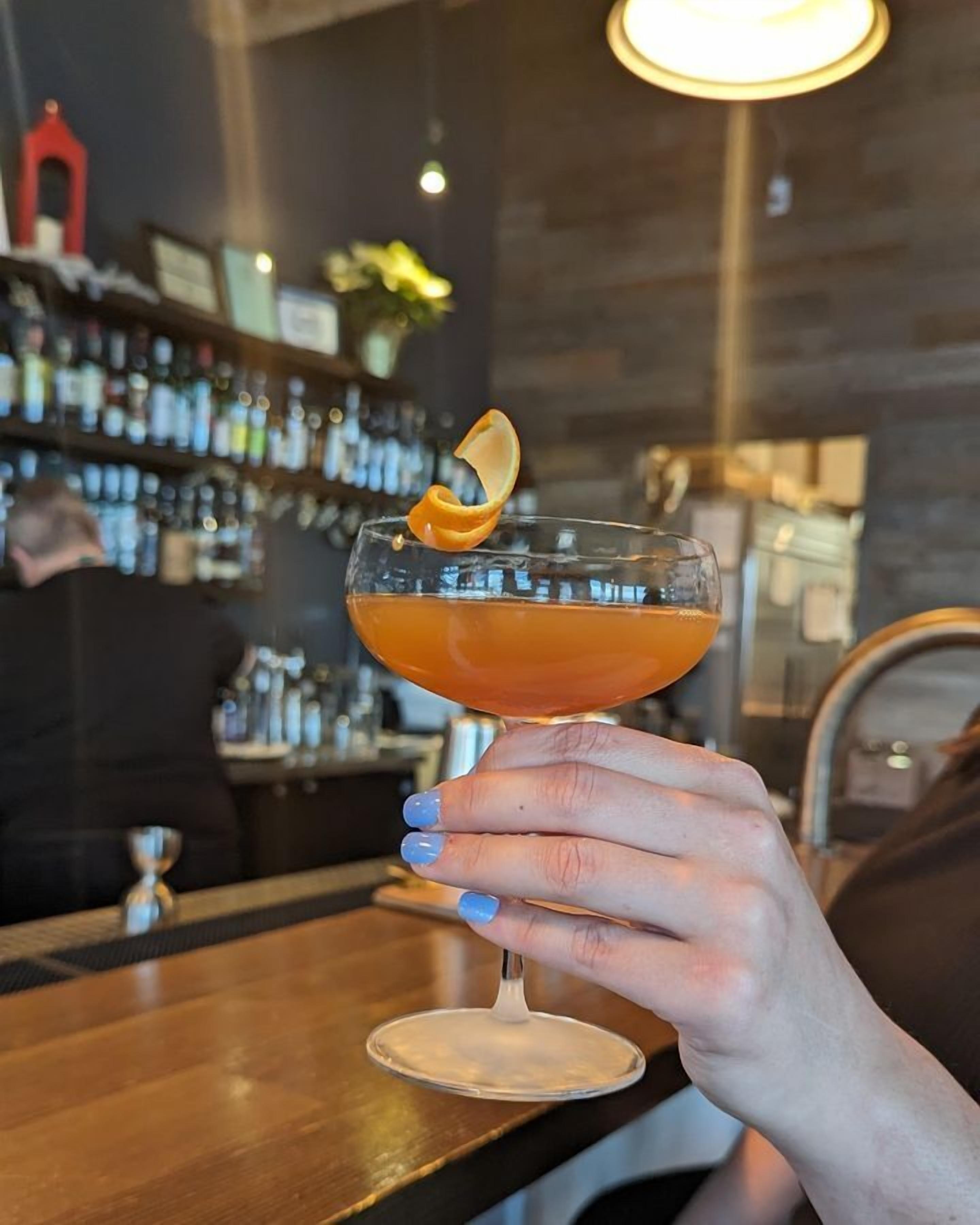 A hand holding a cocktail glass with a citrus garnish, in a bar setting with shelves of bottles and a person in the background.