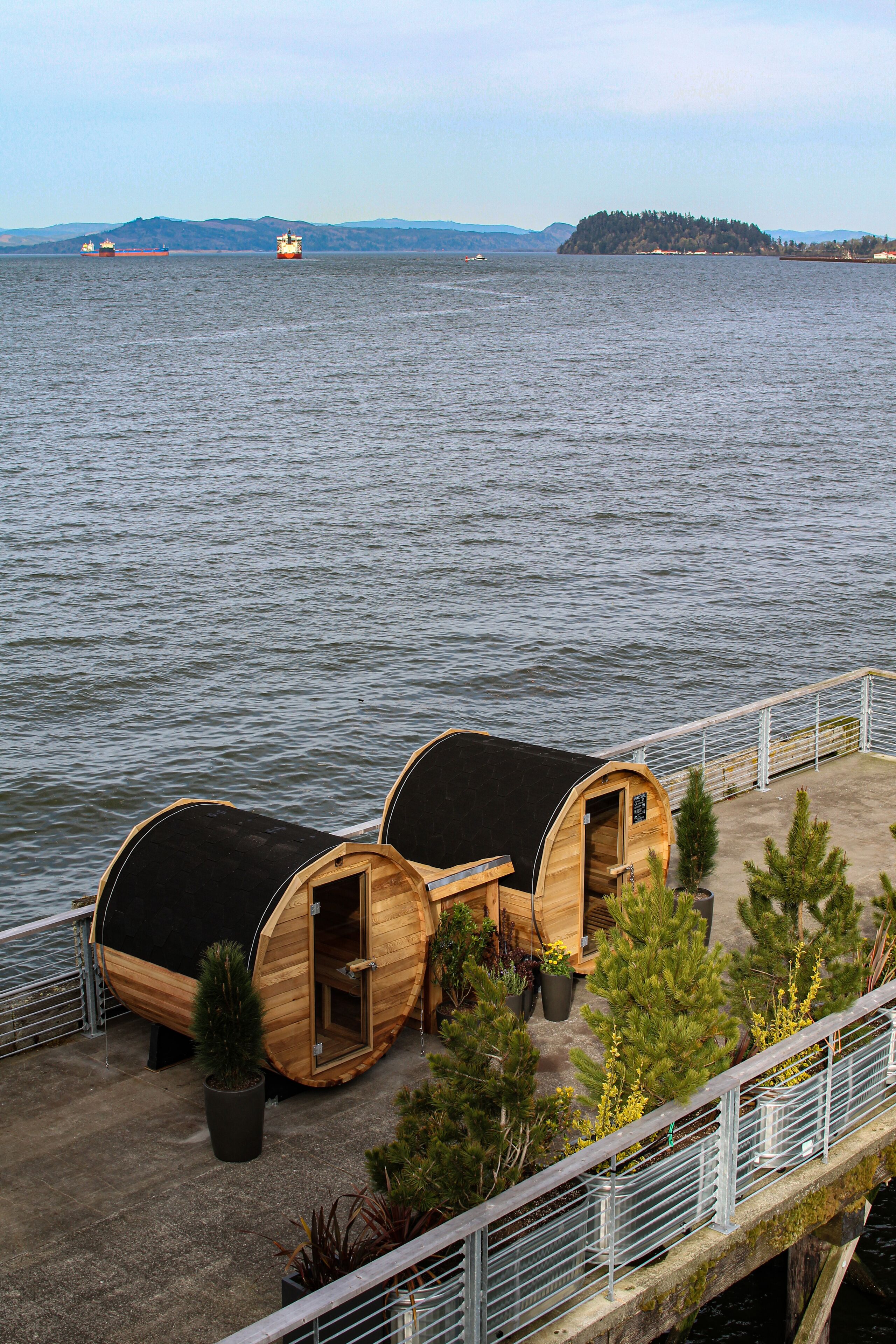 Two wooden barrel-shaped cabins with black roofs, a concrete deck, and a metal railing overlooking a body of water.