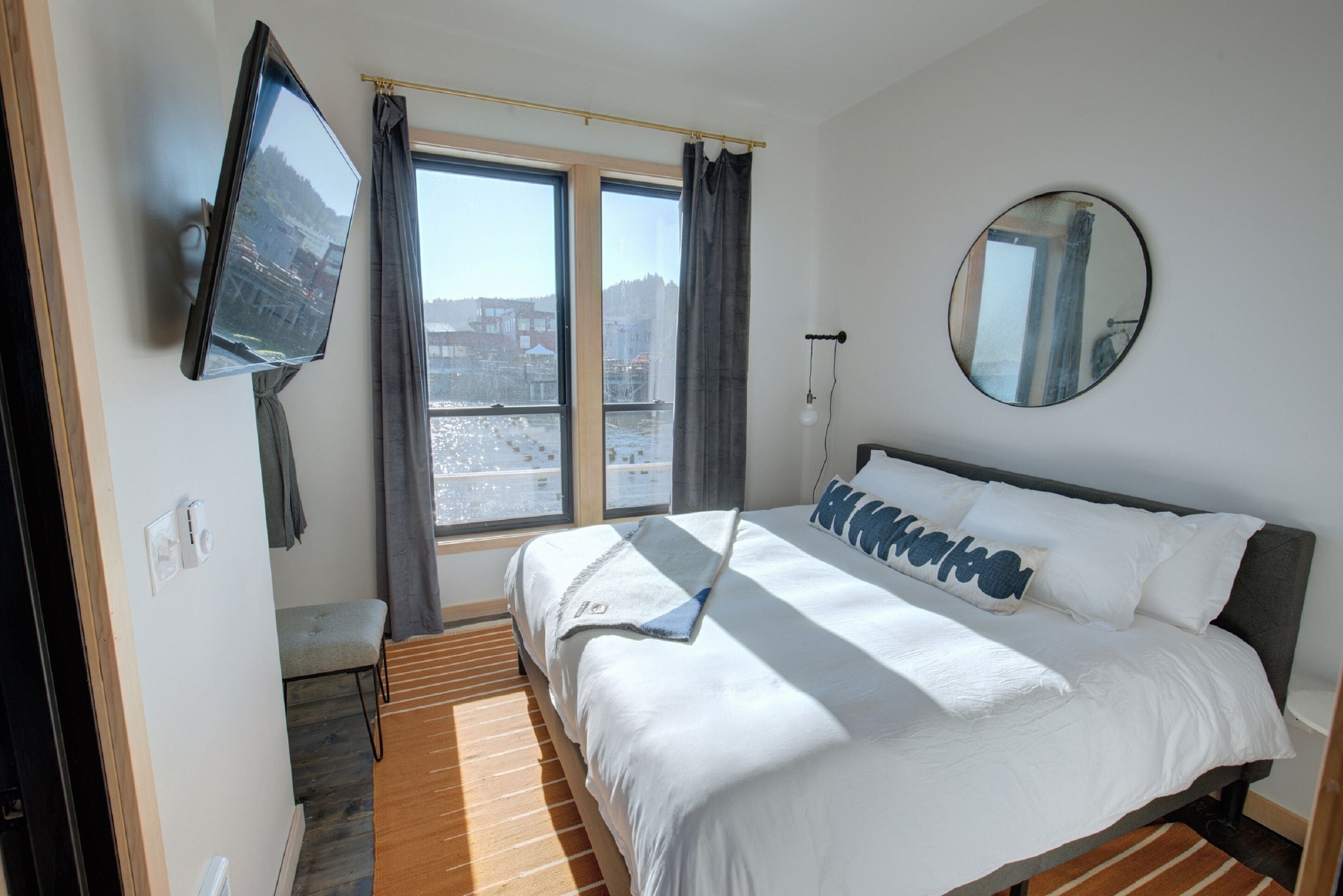 Quarto de hotel com cama, televisão fixada na parede, janela com cortinas, mesa de cabeceira e espelho.