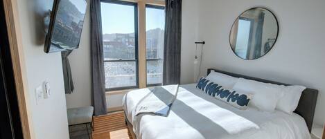 Quarto de hotel com cama, televisão fixada na parede, janela com cortinas, mesa de cabeceira e espelho.