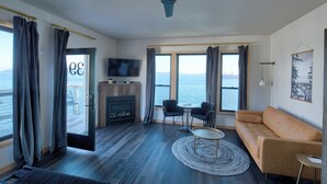 Een moderne woonkamer met een bank, twee fauteuils, een televisie, een open haard en grote ramen met uitzicht op zee.