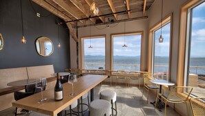 Een restaurant met een houten bar, ronde krukken en grote ramen met uitzicht op zee.